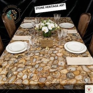 Puede incluir: Una mesa de comedor con una superficie &uacute;nica de piedra de &aacute;gata, preparada para una comida. La mesa est&aacute; adornada con platos blancos, copas de cristal y cubiertos dorados. Tambi&eacute;n se ve un arreglo floral y el texto "STONE HERITAGE ART".