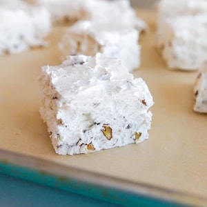 May include: Close-up of several square-shaped, white marshmallows with visible nuts. The marshmallows are on a light brown surface, with a few more marshmallows slightly out of focus in the background. The marshmallows have a textured, slightly uneven surface.