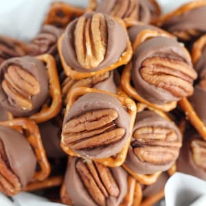 May include: Close-up of chocolate-covered pretzels topped with pecan halves. The pretzels are square-shaped and have a golden-brown color. The chocolate coating is a rich brown, and the pecans are a light brown. The treats are in a white bowl.