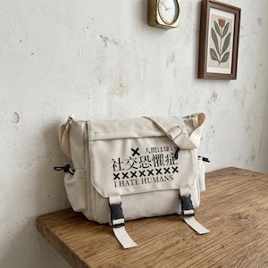 Puede incluir: Un bolso tipo mensajero de lona color crema con un estampado gráfico negro en la solapa frontal. El texto dice "I HATE HUMANS" en inglés y japonés, junto a un símbolo "X" y una fila de "X". Cuenta con dos cierres de hebilla de plástico negro.