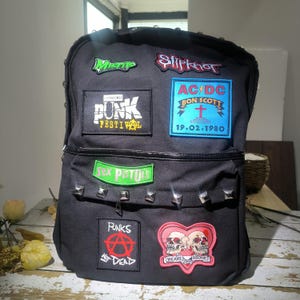 May include: A black backpack decorated with various music-themed patches including Misfits, Slipknot, AC/DC, Sex Pistols, and Punks Not Dead. The bag features a row of silver-toned pyramid studs across the front pocket and a heart-shaped skull patch.