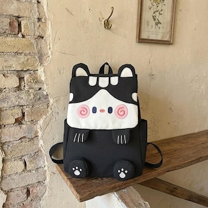 以下が含まれることがあります： フロントフラップに漫画の動物の顔が描かれた黒と白のバックパック。上部に2つの小さな耳、ピンクの渦巻き状の頬、小さな鼻、フラップの下に2つの黒い前足、底面に2つの肉球のプリントがデザインされています。
