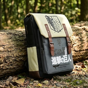 May include: A black and cream-colored backpack featuring the Attack on Titan logo and Japanese text on the front flap and main body. The bag includes two brown faux leather straps with metal buckles, a side pocket, and a top flap closure design.