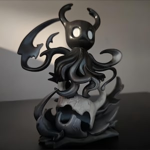 May include: A dark gray figurine of a character with a horned head, large eyes, and flowing cape-like appendages. The figure is mounted on a stylized base with a skull and flame-like shapes. The overall design is detailed and artistic.