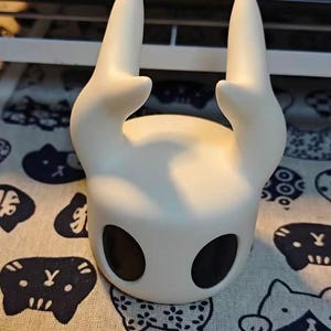 Puede incluir: Una escultura blanca de la cabeza de un personaje con grandes cuencas de ojos negros y dos cuernos curvos. El objeto liso y mate tiene una forma redondeada. Se coloca sobre una tela estampada con figuras negras parecidas a gatos.