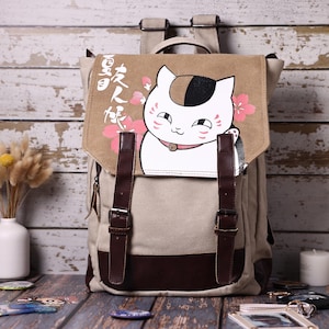 Op de afbeelding: Een beige canvas rugzak met een flap voorzien van een afbeelding van een witte, zwarte en oranje kat, roze kersenbloesems en witte Japanse tekst. De tas heeft donkerbruine kunstlederen riemen met metalen gespen en een donkerbruin basispaneel.
