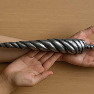 May include: A silver-coloured, spiralled horn-shaped object. The horn has a textured, twisted design and tapers to a sharp point. The object is held in two hands, with a wooden surface in the background.
