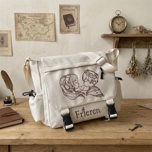 May include: A cream-coloured canvas messenger bag featuring a brown line-art illustration of two anime-style characters connected by a heart string. The bag has a flap with two black plastic buckles and the text Frieren printed in brown.