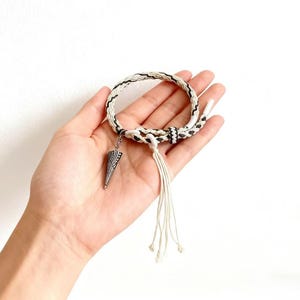 Op de afbeelding: Een handgemaakte armband met een geweven ontwerp in zwart en wit, met een zilverkleurige hanger. De armband heeft een kwastje van witte koorden en wordt in een hand tegen een witte achtergrond gehouden. Een stijlvol accessoire.