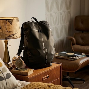 May include: A black backpack with white text, placed on a wooden bedside table. An alarm clock and a book are beside the bag. A lamp with a patterned shade and a brown leather chair are also in the room.