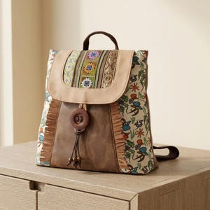 May include: A brown and beige backpack with a decorative flap featuring embroidered patterns. The backpack has a top handle, a large wooden button closure, and a patterned fabric side with fringe detailing. The bag is sitting on a wooden surface.