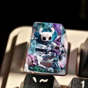 May include: A custom artisan keycap with a white horned character in a clear resin keycap. The design includes blue, purple, and pink details, forming a small scene. The keycap is on a keyboard.