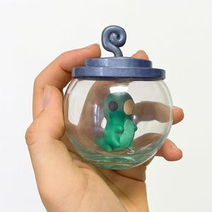 Pode incluir: Um frasco de vidro transparente com tampa azul escuro e uma pega decorativa em espiral. Dentro do frasco está uma pequena criatura verde, semelhante a um desenho animado, com um rosto preto e uma mancha bege. O frasco é segurado na mão.