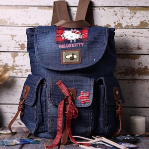 Puede incluir: Una mochila de mezclilla azul oscuro con un gráfico de Hello Kitty sobre un parche de cuadros rojos, el texto HELLO KITTY, un cierre de metal, correas marrones, dos bolsillos frontales con detalles marrones y un lazo de cuadros rojos.