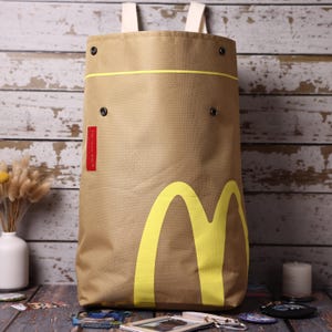 Puede incluir: Una mochila de color tostado con un gran gráfico amarillo en forma de M en la parte delantera, una línea amarilla horizontal y cuatro botones de presión metálicos negros. Una pequeña etiqueta rectangular roja en el lado izquierdo muestra el texto blanco I'm lovin' it. Tiene correas blancas.