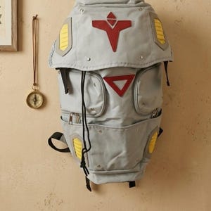 以下が含まれることがあります： 赤と黄色のアクセントが特徴的なグレーのバックパック。ユニークなデザインで、複数のポケット、巾着開閉、フラップが付いています。装飾的なリベットとパッチが特徴的な外観を添えています。バッグは壁に掛けられています。