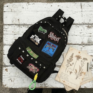 May include: Black backpack adorned with patches of bands such as Misfits, Slipknot, and AC/DC. The bag features silver studs and a green and yellow keychain. A vintage botanical print is placed on the side.