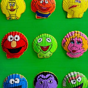 May include: Nine painted seashell art pieces featuring Sesame Street and Muppets characters on a green background. Characters include Bert, Ernie, Big Bird, Elmo, Kermit, Miss Piggy, Cookie Monster, The Count, and a green monster.