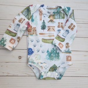 Op de afbeelding: Een witte baby romper met lange mouwen met een campingthema. De print bevat verschillende kampeerartikelen, waaronder tenten, kampvuren, bomen en een kano. De romper heeft een drukknoopsluiting aan de onderkant.
