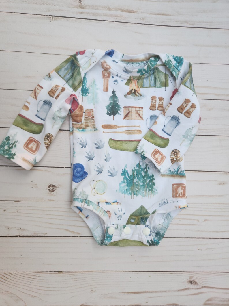 Op de afbeelding: Een witte baby romper met lange mouwen met een campingthema. De print bevat verschillende kampeerartikelen, waaronder tenten, kampvuren, bomen en een kano. De romper heeft een drukknoopsluiting aan de onderkant.