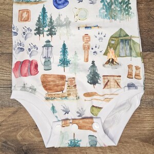 Op de afbeelding: Witte baby romper met een aquarelprint van campingthema's, waaronder bomen, een tent, een lantaarn, een slaapzak, een kampvuur en een kompas.