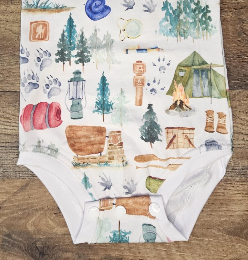 Op de afbeelding: Witte baby romper met een aquarelprint van campingthema's, waaronder bomen, een tent, een lantaarn, een slaapzak, een kampvuur en een kompas.