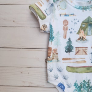 Op de afbeelding: Een witte baby romper met een campingthema print. De print bevat verschillende kampeerartikelen, waaronder een tent, een kampvuur, een kano en een kompas. De romper heeft korte mouwen en drukknopen aan de onderkant.