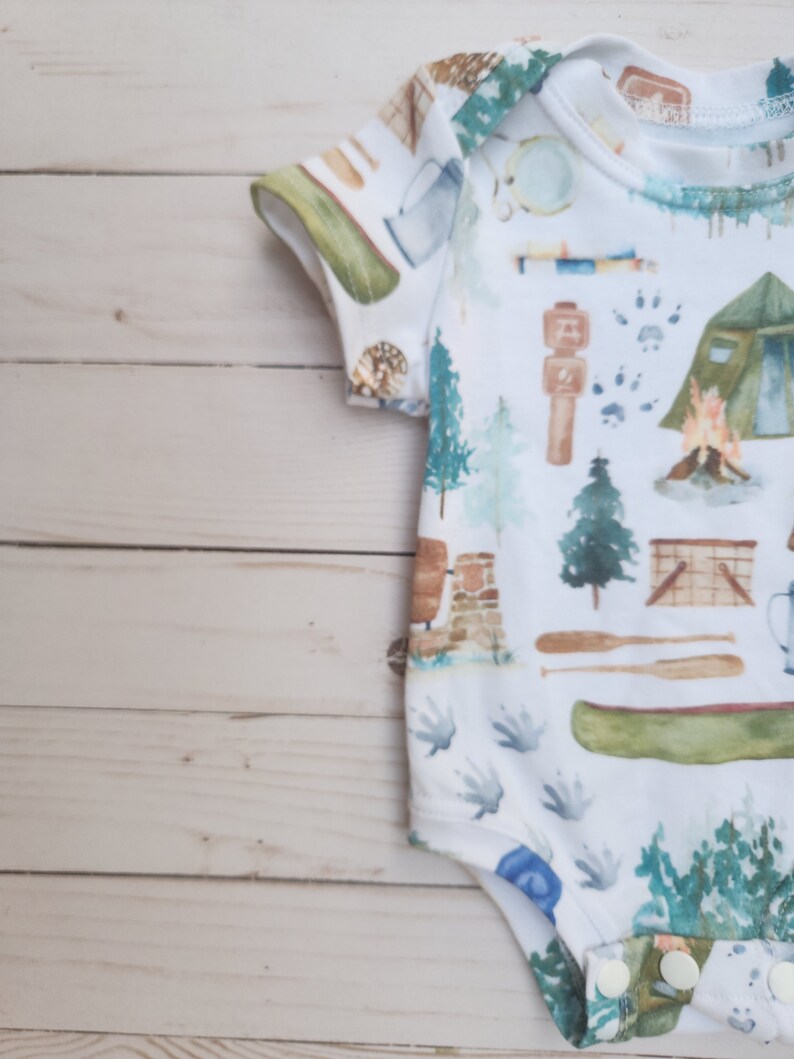 Op de afbeelding: Een witte baby romper met een campingthema print. De print bevat verschillende kampeerartikelen, waaronder een tent, een kampvuur, een kano en een kompas. De romper heeft korte mouwen en drukknopen aan de onderkant.