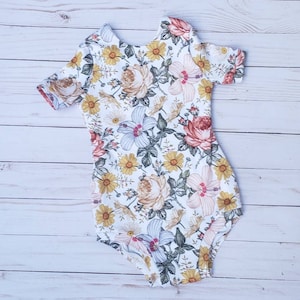 May include: A white bodysuit with a floral print featuring yellow, pink, red, and green flowers. The bodysuit has short sleeves and a round neckline.