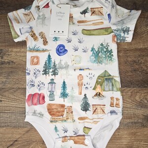 Op de afbeelding: Een witte baby romper met een aquarelprint van kampeeruitrusting, waaronder een tent, een kano, een lantaarn, een slaapzak en een kampvuur. De romper heeft korte mouwen en drukknoopjes aan de onderkant. De tekst "Soft Bling Design" staat op een label dat aan de romper is bevestigd.