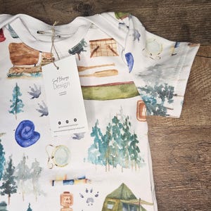Op de afbeelding: Witte baby romper met een aquarelprint van kampeeruitrusting, waaronder een tent, kampvuur, bomen en een kompas. De romper heeft een label met de tekst "Soul Blessings Design Clothing Company".