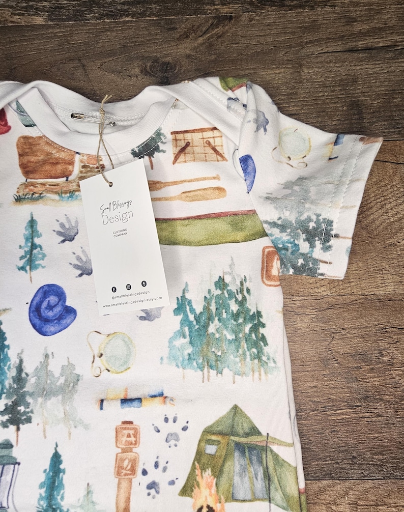 Op de afbeelding: Witte baby romper met een aquarelprint van kampeeruitrusting, waaronder een tent, kampvuur, bomen en een kompas. De romper heeft een label met de tekst "Soul Blessings Design Clothing Company".