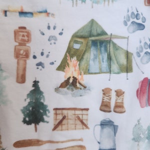 Op de afbeelding: Een witte stof met een herhalend patroon van camping- en natuurthema illustraties. Het patroon omvat een tent, een kampvuur, een kano, een picknickmand, een koffiekan, een paar laarzen, een boom, dierenpootjes en een bord met een kampvuur symbool.