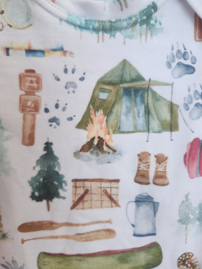 Op de afbeelding: Een witte stof met een herhalend patroon van camping- en natuurthema illustraties. Het patroon omvat een tent, een kampvuur, een kano, een picknickmand, een koffiekan, een paar laarzen, een boom, dierenpootjes en een bord met een kampvuur symbool.