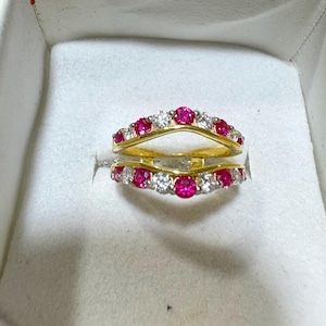 May include: A gold-toned ring with a curved design, set with alternating red and clear gemstones. The ring is displayed inside a white jewelry box, showcasing its intricate details and vibrant colors.
