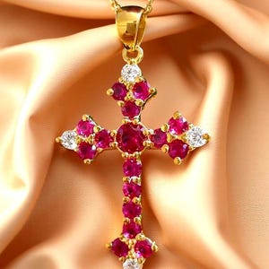 Può includere: Una collana con pendente a forma di croce color oro con una catena. La croce è adornata da numerose pietre preziose rosse rotonde e a forma di pera, e pietre preziose bianche. La collana è esposta su un tessuto color pesca.
