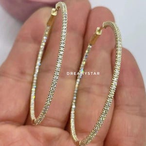 Könnte beinhalten: Ein Paar goldene Creolen, besetzt mit kleinen, funkelnden Edelsteinen. Die Ohrringe sind oval geformt und weisen eine durchgehende Reihe klarer Steine auf. Das Bild ist gut beleuchtet und unterstreicht die Brillanz der Edelsteine. Die Ohrringe werden von einer Hand gehalten.
