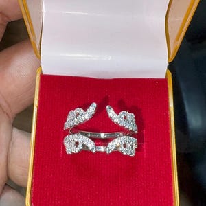 Puede incluir: Un anillo de plata con un diseño de doble banda, adornado con numerosas piedras preciosas pequeñas y transparentes. El anillo se presenta en una caja forrada de terciopelo rojo con exterior amarillo.