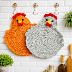 Puede incluir: Dos salvamanteles de ganchillo hechos a mano con forma de gallina. Uno es naranja, el otro gris. Ambos tienen crestas rojas, picos amarillos y ojos negros. Cuelgan de ganchos dorados contra una pared de azulejos blancos.