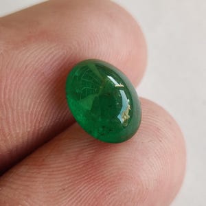 May include: A vibrant, oval-shaped emerald gemstone. The polished surface reflects light, showcasing a deep green hue. The gemstone is held between fingers, highlighting its size and clarity. This gemstone is ideal for jewelry making.