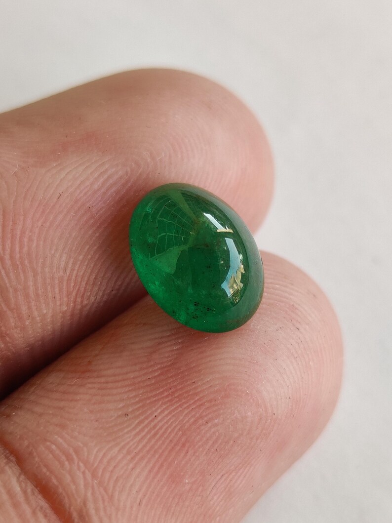 May include: A vibrant, oval-shaped emerald gemstone. The polished surface reflects light, showcasing a deep green hue. The gemstone is held between fingers, highlighting its size and clarity. This gemstone is ideal for jewelry making.