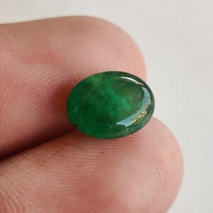 May include: A vibrant, oval-shaped emerald gemstone. The polished surface reflects light, showcasing the rich green color and internal patterns. The gemstone is held between fingers, highlighting its size and shape.