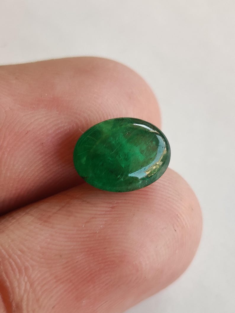May include: A vibrant, oval-shaped emerald gemstone. The polished surface reflects light, showcasing the rich green color and internal patterns. The gemstone is held between fingers, highlighting its size and shape.