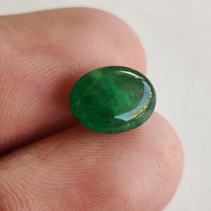 May include: A vibrant, oval-shaped emerald gemstone. The polished surface reflects light, showcasing a deep green color with subtle internal patterns. The gem is held between fingers, highlighting its size and clarity.
