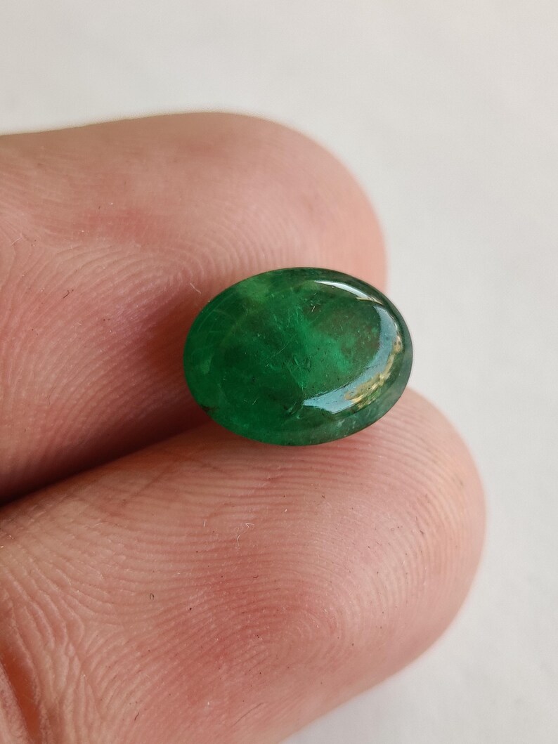 May include: A vibrant, oval-shaped emerald gemstone. The polished surface reflects light, showcasing a deep green color with subtle internal patterns. The gem is held between fingers, highlighting its size and clarity.