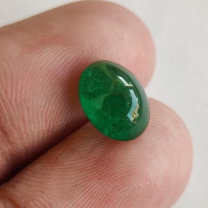 May include: A vibrant, oval-shaped emerald gemstone. The polished surface reflects light, showcasing a deep green hue. The gem is held between fingers, highlighting its size and clarity. This gemstone is suitable for jewelry making.