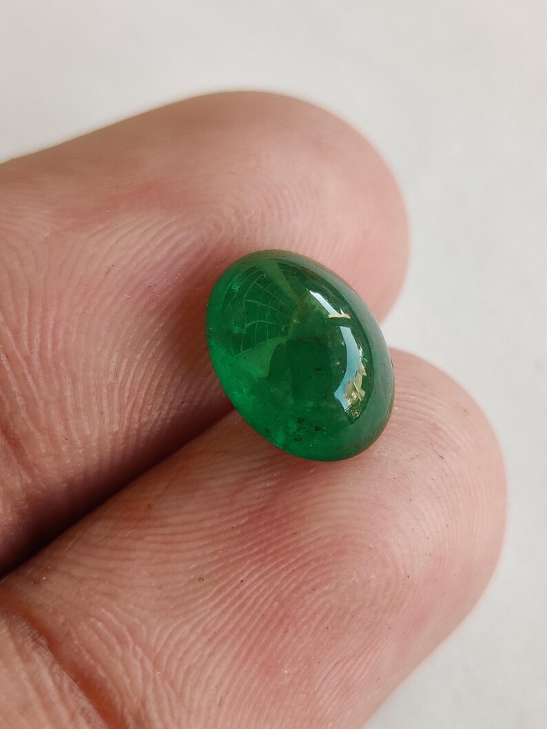 May include: A vibrant, oval-shaped emerald gemstone. The polished surface reflects light, showcasing a deep green hue. The gem is held between fingers, highlighting its size and clarity. This gemstone is suitable for jewelry making.