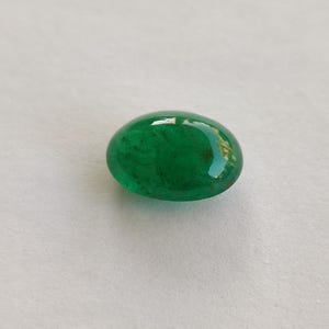 May include: A vibrant, oval-shaped emerald gemstone. The polished surface reflects light, showcasing a deep, rich green color. The gemstone is set against a neutral background, highlighting its natural beauty. This gemstone is ideal for jewelry making.