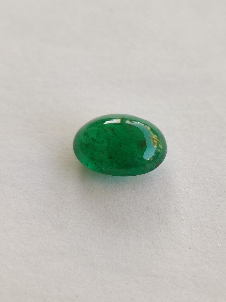 May include: A vibrant, oval-shaped emerald gemstone. The polished surface reflects light, showcasing a deep, rich green color. The gemstone is set against a neutral background, highlighting its natural beauty. This gemstone is ideal for jewelry making.