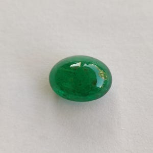 May include: A vibrant, oval-shaped, polished green gemstone. The gem has a smooth, glossy surface, reflecting light. The color is a rich, deep green, with subtle variations in tone. The gemstone is set against a plain, light background.